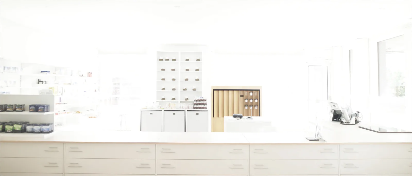 The interior of an independent pharmacy in daylight — white shelving, a drawered counter, and a wooden dispensing unit, with no one in the room